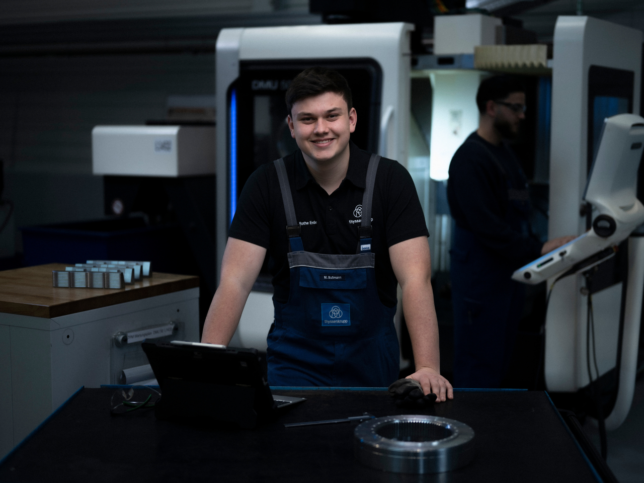 Junger Mann vor einem Laptop, hinter ihm eine CNC-Drehmaschine mit Bildschirm auf der rechten Seite.