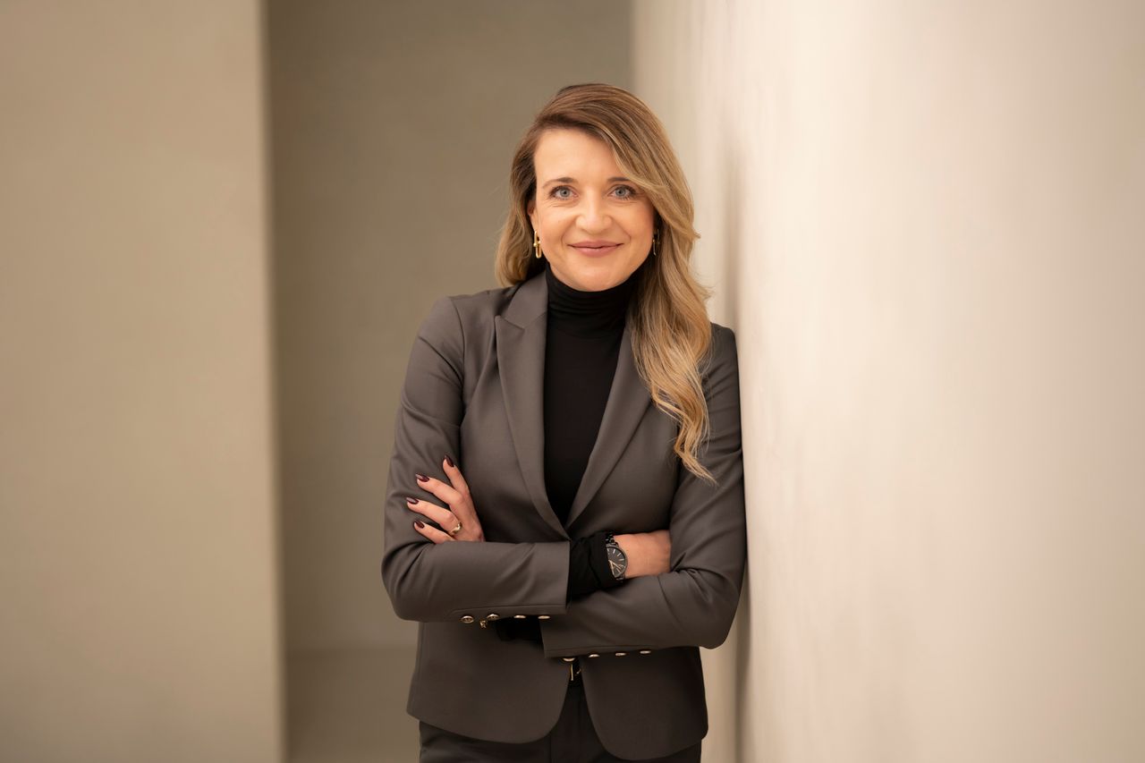 Blonde woman, smiling, arms crossed, wearing a black shirt and a grey blazer