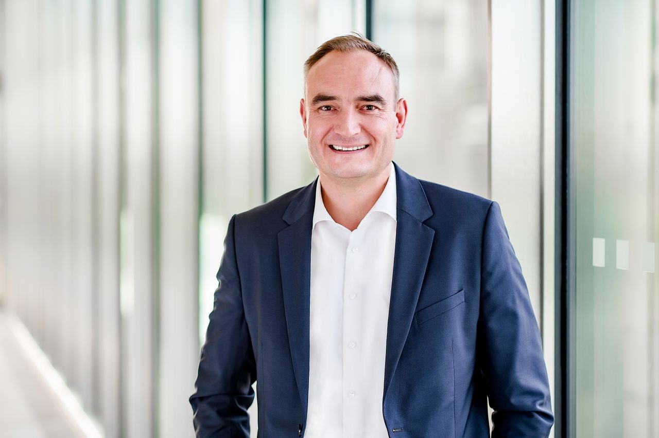 Homme souriant, en chemise et veste devant un mur de verre