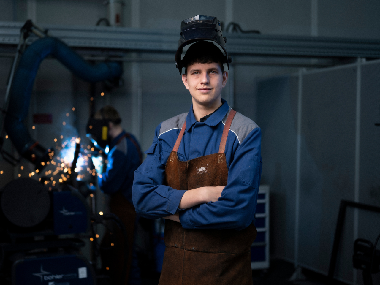 Junger Mann mit Schweißhelm auf dem Kopf, seine Arme sind verschränkt. Im Hintergrund eine Person am schweißen.