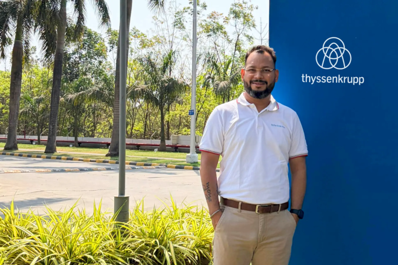 A person stands outdoors next to a blue thyssenkrupp sign, wearing a polo shirt and trousers, with trees and greenery visible in the background.
